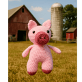 Crocheted pink pig toy in a field with a barn and silo in the background