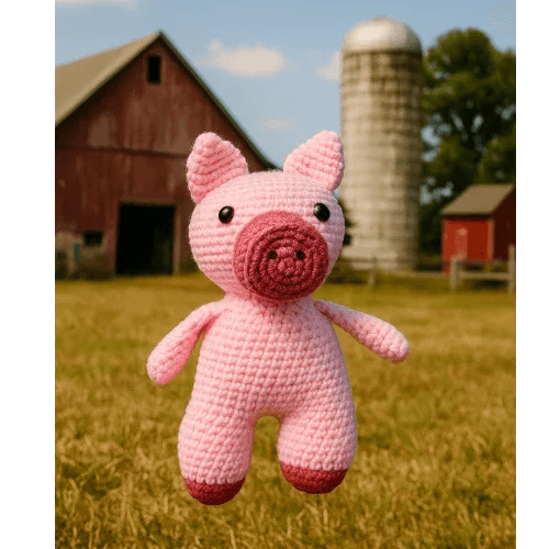 Crocheted pink pig toy in a field with a barn and silo in the background