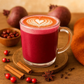 Glass mug of red beet juice with a heart foam design, surrounded by pomegranates, cinnamon sticks, and star anise on a wooden surface.