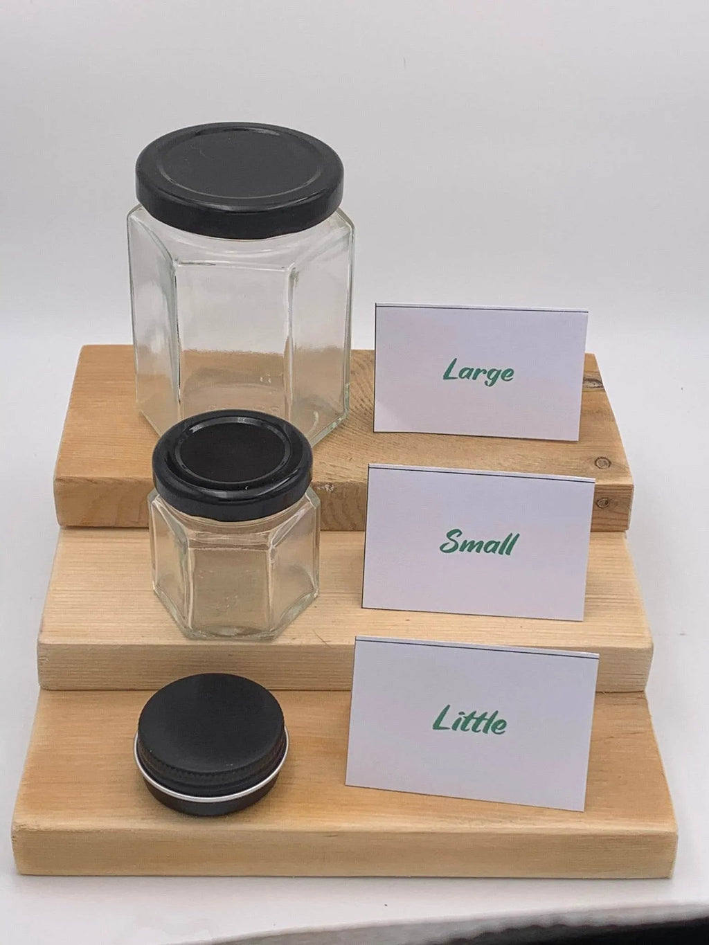 Three hexagonal glass jars with black lids on a wooden stand, labeled 'Large', 'Small', and 'Little'.
